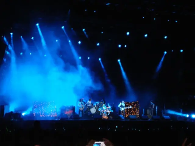 In this image on the stage few people are performing. On the top there are lights. In the...